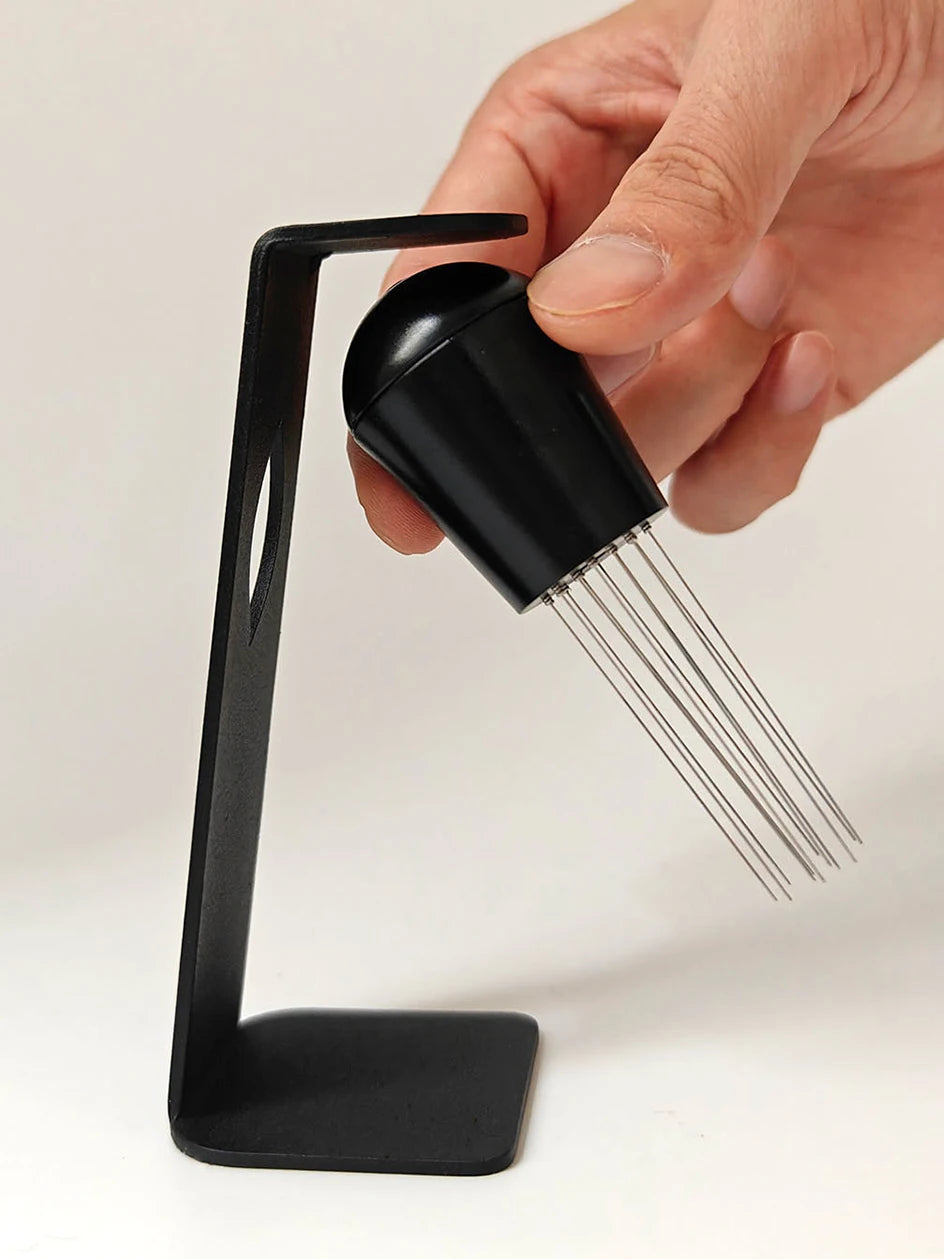 Hand holding espresso distribution tool with fine needles beside black metal stand, coffee accessory