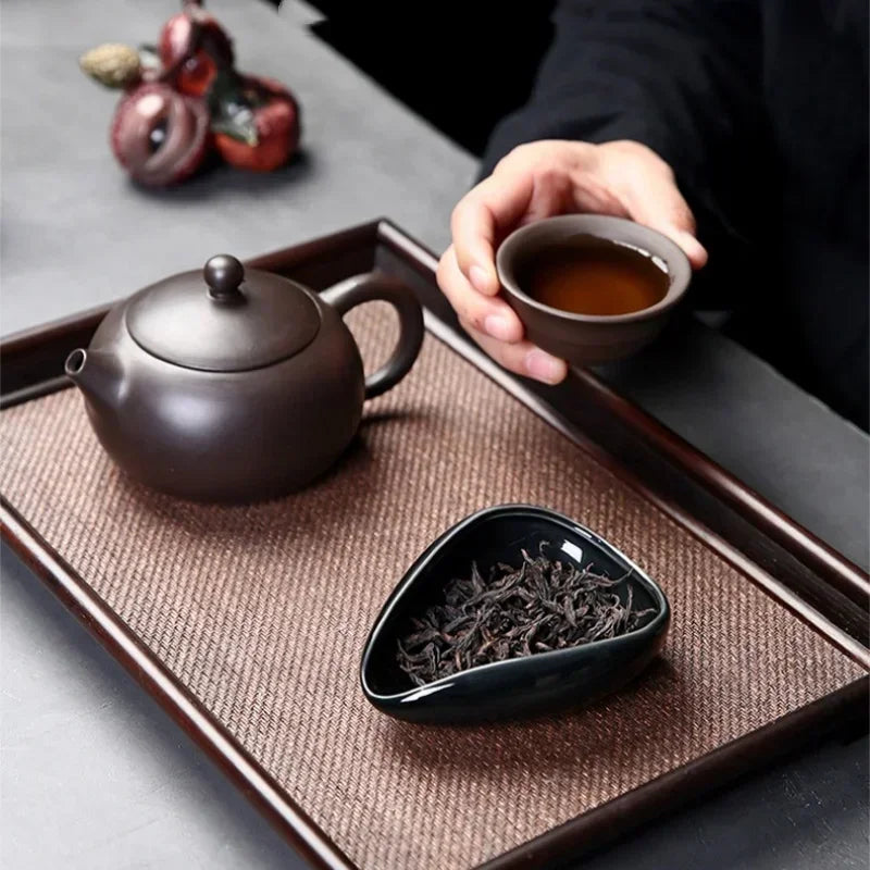 Traditional tea set with dark teapot, loose leaf tea, and hand holding teacup on tray.